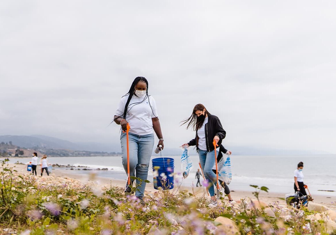 GRANTEE-Watershed-Protection-Fund_-Santa-Barbara-Waterkeeper A group of young people, including a Black woman with long braids, and a while woman with long brown hair, wearing masks and pikcing up garbage with orange grabbers along a coastline on a grey day.