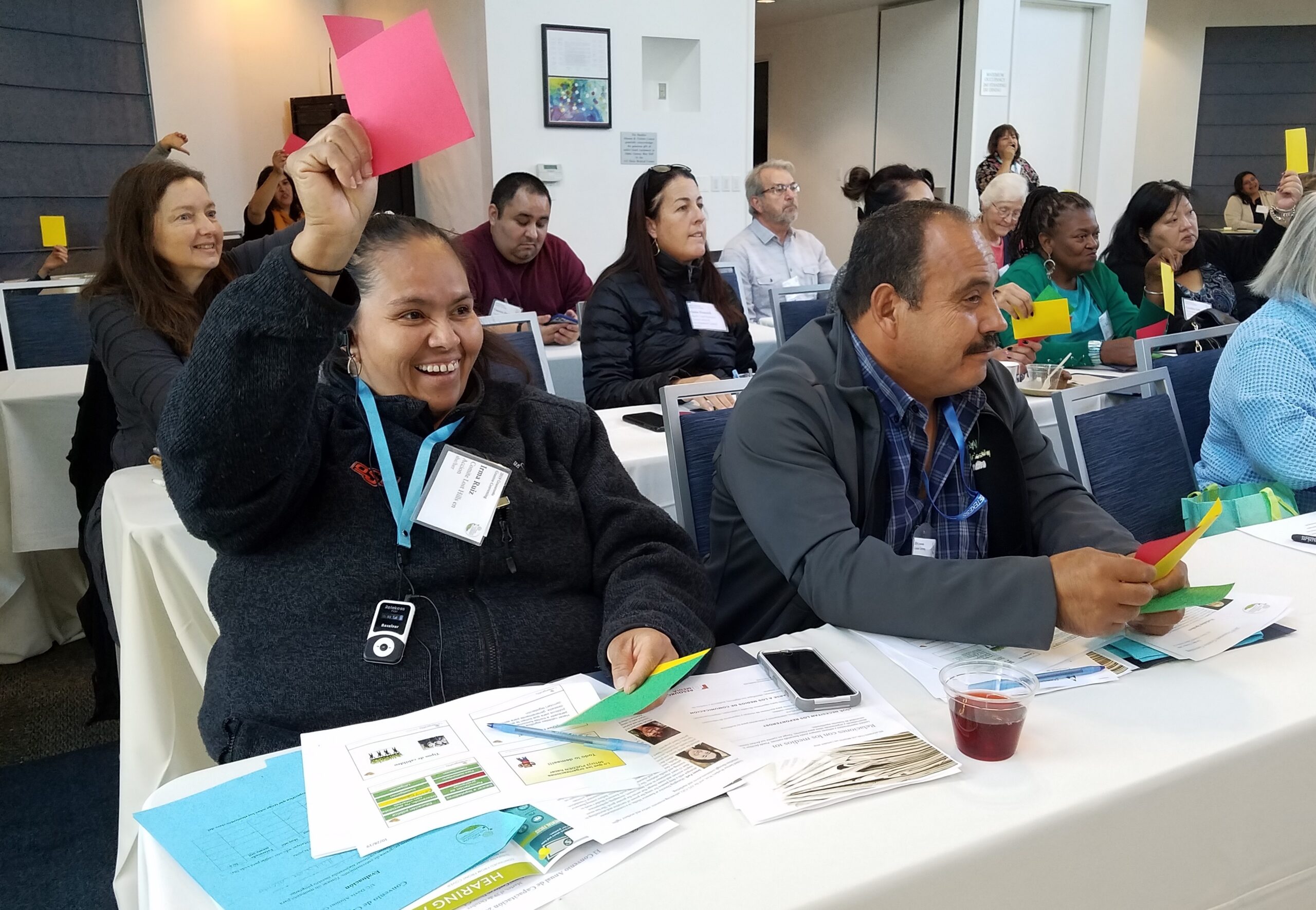 A diverse group of adults sit in a conference room. Some raise colored cards; a smiling woman in front holds up a red card and a man next to her holds a yellow one. Papers and a cup with a drink are on the table. Everyone appears engaged and attentive.