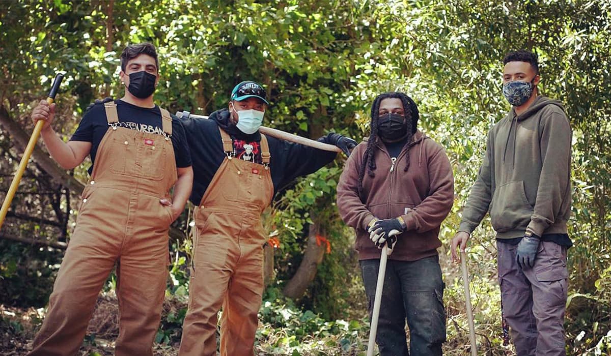 4 young men of various races, wearing overalls and carying shovels, posing in a forested setting.